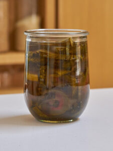 Glass jar filled with pickled watermelon rind pieces in dark amber brine, resting on a white surface. The rind strips appear translucent olive green with a few pink edges visible through the glass.