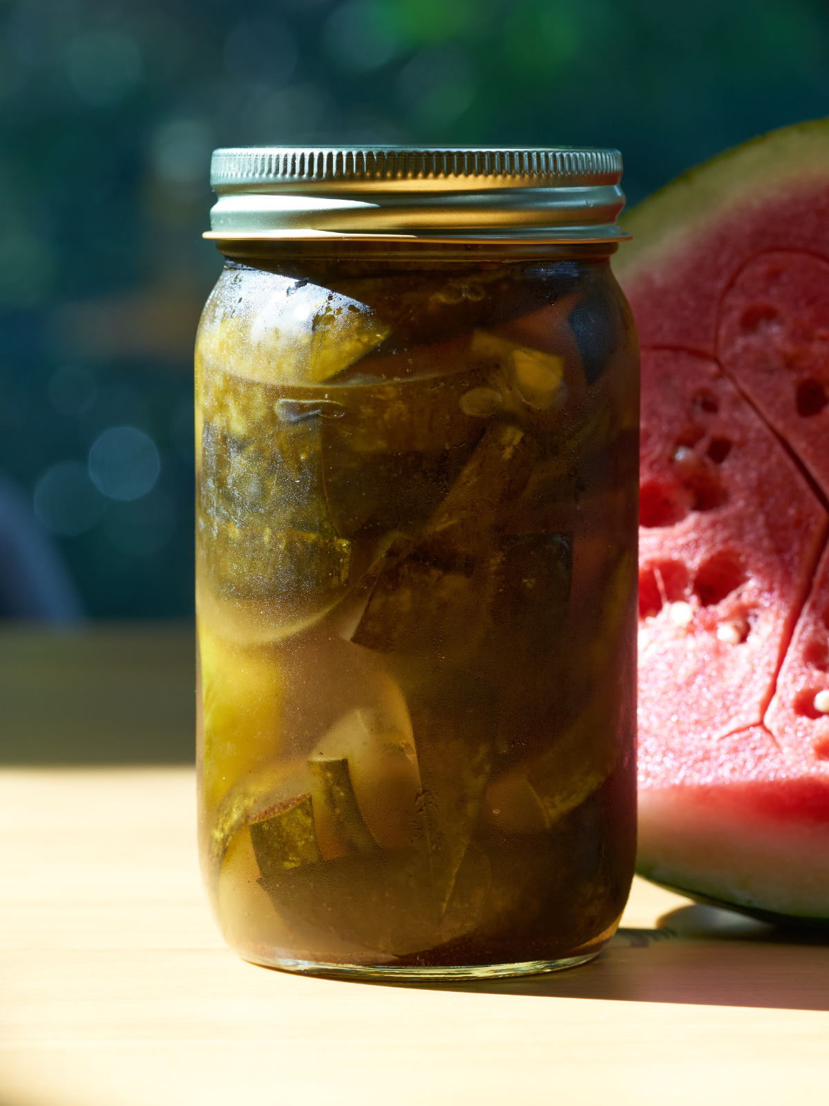 Jar of green pickled watermelon rind next to half a watermelon on a wood table.