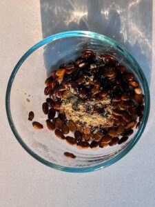 A clear glass bowl filled with dark roasted watermelon seeds topped with light brown ground spice.
