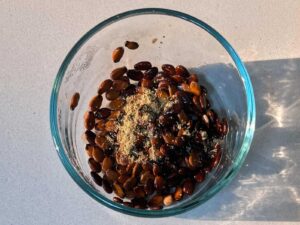 A clear glass bowl filled with dark roasted watermelon seeds topped with light brown ground spice.