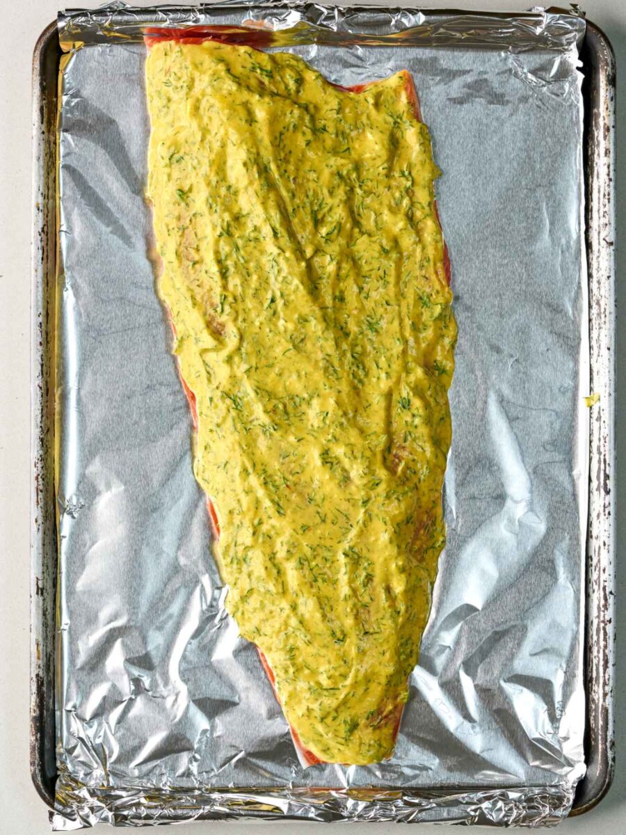 A raw, whole salmon side on a foil-lined baking sheet covered with a thick layer of yellowish herb sauce.