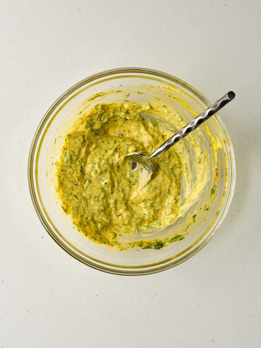 A mixing bowl containing thick yellow yogurt sauce mixed with specks of fresh chopped dill.